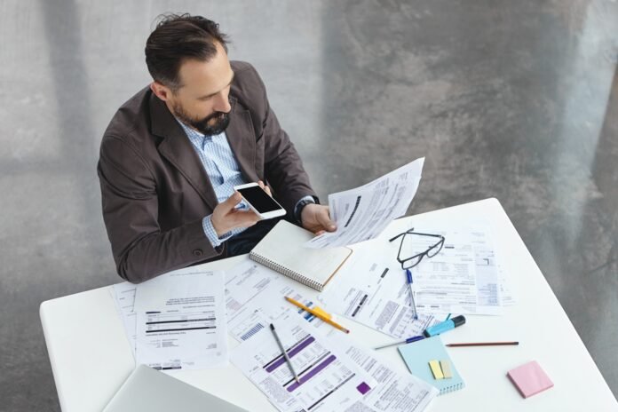 Portrait of busy male office worker makes voice call, discuss documents with partner, compare result, try to predict wastes for next month. Head executive officer holds balance sheet and mobile phone